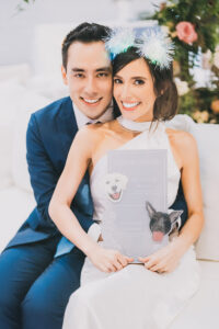 Smiling couple at Gulf South wedding; woman in white dress holds dog-themed cocktail sign, man wears blue suit. Flowers blurred behind.