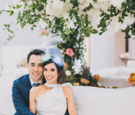 Smiling couple in formalwear on a white couch at their New Orleans wedding, with white flowers and a blue headband.