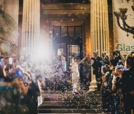 Newlyweds walk down grand steps at night as guests blow bubbles and take photos, celebrating a lively New Orleans wedding.