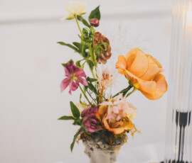 Head-shaped vase with orange, pink, and yellow flowers next to a lit candle and ribbed lamp on a white surface.