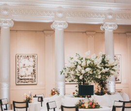 Grand hall wedding setup with white columns, round tables, black chairs, floral centerpieces, and framed art on cream walls.
