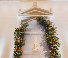 White tiered cake with floral decor framed by an orange and yellow flower arch, set before ornate molding at a Louisiana wedding.