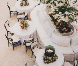 Round tables with white linens, floral centerpieces, and green candles near white couches in a bright Gulf South wedding venue.