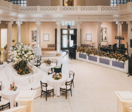 Spacious New Orleans event hall with round tables, floral centerpieces, white columns, and a stage—perfect for weddings.