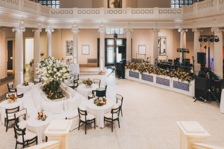 Spacious New Orleans event hall with round tables, floral centerpieces, white columns, and a stage—perfect for weddings.