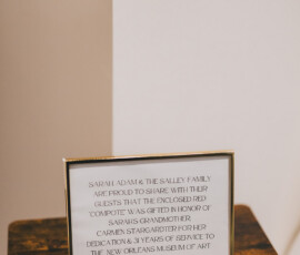Framed sign on wood highlights a red compote gift honoring Carmen Strapp’s 31 years at New Orleans Museum of Art.