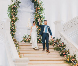 Bride in white dress and groom in blue suit walk down floral stairs, holding hands and smiling after their wedding.
