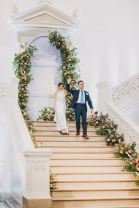 Bride in white dress and groom in blue suit walk down floral stairs, holding hands and smiling after their wedding.