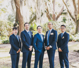 Five men in blue suits pose outdoors with mossy trees and sunlight, perfect for New Orleans wedding inspiration.