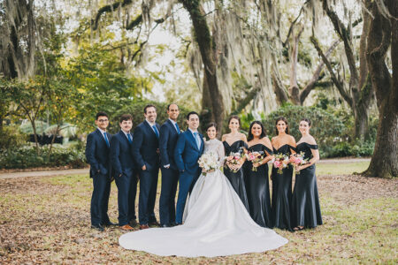 Bride and groom with bridesmaids in black and groomsmen in blue pose outdoors under mossy trees at a Louisiana wedding.