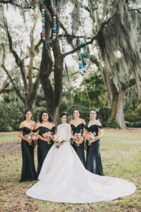 Bride in white gown with flowers stands by four bridesmaids in black, outdoors with trees and vibrant hanging wedding ornaments.