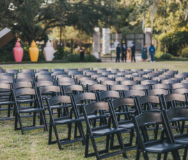 Outdoor Louisiana wedding setup with black folding chairs on grass, colorful vases, trees, and people in the background.