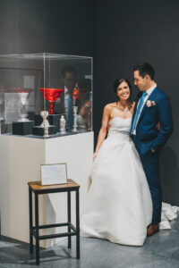 Bride in white gown and groom in blue suit smile beside ornate goblets at their Louisiana wedding in a museum setting.