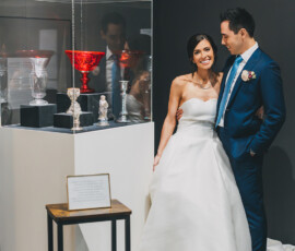 Bride in white gown and groom in blue suit smile beside ornate goblets at their Louisiana wedding in a museum setting.