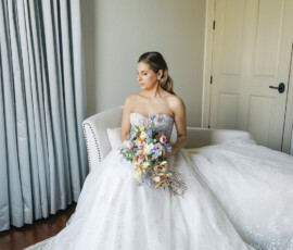Bride seated on a cream sofa holding a colorful garden-style bouquet, wearing a strapless ball gown in a softly lit room.