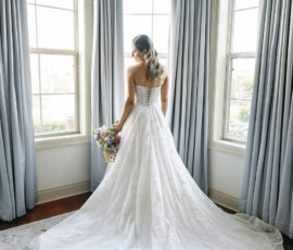 Back view of the bride standing between tall windows, showcasing the flowing train of her wedding gown and pastel bouquet.