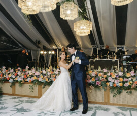 Bride and groom sharing their first dance beneath woven pendant lights and a lush floral stage backdrop inside the reception tent.