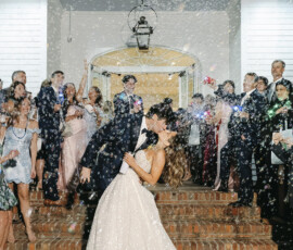 Bride and groom sharing a dramatic dip kiss during their sparkler-style bubble exit, surrounded by cheering guests on the venue steps at night.