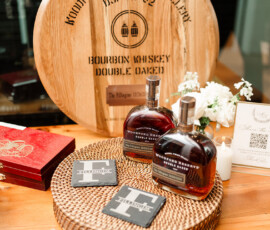 Woodford Reserve bourbon bottles on a wicker tray with coasters, candle, flower, red box, and engraved barrel top—ideal for beach weddings.
