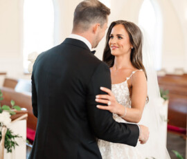 Bride in white dress and veil and groom in black tuxedo share a loving gaze at their Orange Beach church wedding.
