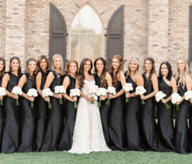 Bride in white gown with twelve bridesmaids in black dresses, holding white bouquets outside brick building before Gulf Shores wedding.