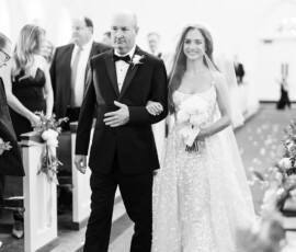 Bride in floral gown and veil walks down church aisle with older man; flower petals decorate the floor at a classic Gulf Shores wedding.