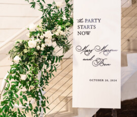 Chic white wedding sign with greenery reads: The party starts now. Mazi Mazure & Ben, October 26, 2024.
