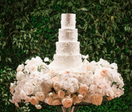 Elegant five-tier white wedding cake with floral accents on glass stand, surrounded by blooms and green foliage in Alabama.
