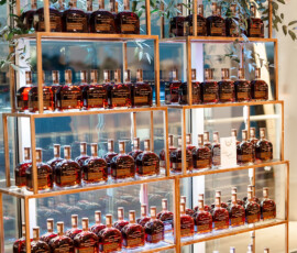 Glass bottles with amber liquid on gold shelves, green plants atop, against a glass wall—an elegant Gulf Shores wedding display.
