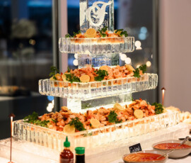 Three-tier ice sculpture with shrimp, garnishes, sauces, and sushi on a buffet by large windows in a softly lit room.