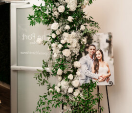 White rose and greenery arrangement by a door, beside a smiling couple’s photo at an elegant Orange Beach wedding event.