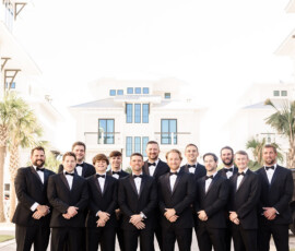Thirteen men in black tuxedos pose outdoors by white buildings and palm trees at an Orange Beach Alabama wedding.