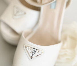 Prada white high heels with ankle straps and metal logo plate, set on a soft background with a blurred white flower.