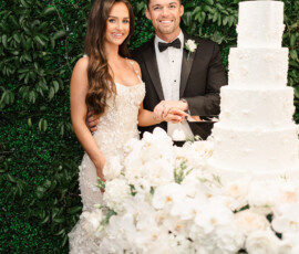 Bride and groom smiling as they cut a white wedding cake at an elegant Orange Beach reception with lush greenery and white flowers.