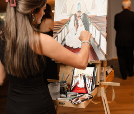 Woman in black dress paints Orange Beach Alabama wedding scene from a photo; canvas shows bride and groom kissing in church.