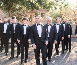 Men in black tuxedos and bow ties smile outdoors by a brick structure at an Orange Beach Alabama wedding.