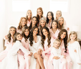 Bride in white robe on bed with smiling bridesmaids in pink pajamas and girls in white nightgowns before Orange Beach wedding.