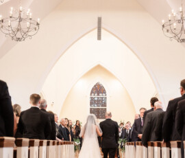Bride in white gown walks down church aisle under chandeliers and stained glass, as guests watch, creating a breathtaking atmosphere.