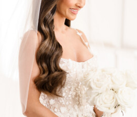 Bride with long wavy brown hair in a white floral dress and veil smiles at her white rose bouquet at Orange Beach wedding.