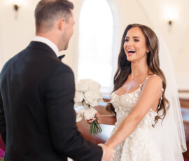 Bride in white dress smiles at groom in black suit, holding white bouquet in a bright Orange Beach wedding chapel.