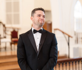 Man in a black tuxedo stands indoors, smiling with hands clasped; elegant Orange Beach wedding reception setting in background.