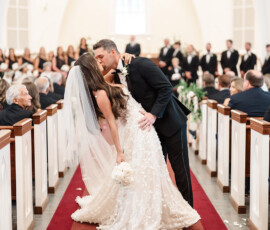 Bride and groom kiss at church aisle end, surrounded by guests and white petals on red carpet at Gulf Shores wedding.