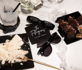 Wedding table at Orange Beach: cake plates, sunglasses, napkin with date, chocolate strawberry, drink, white floral decor.