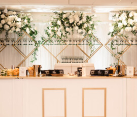 Elegant white and gold bar with floral decor, glassware shelves, and white curtain backdrop for an Orange Beach wedding.
