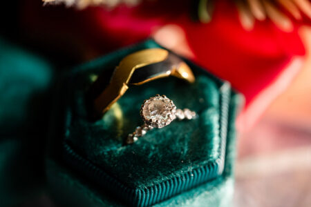 Diamond engagement ring and black-gold wedding band on green velvet box, with blurred red flower in background.