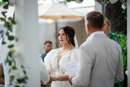 Bride in white dress and veil holds hands with groom in light suit at outdoor Gulf South wedding, lush greenery and flowers around.