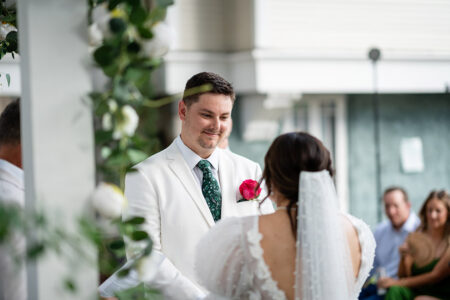 Groom in white suit with pink boutonniere smiles at bride in veil during outdoor Louisiana wedding; guests seated behind.
