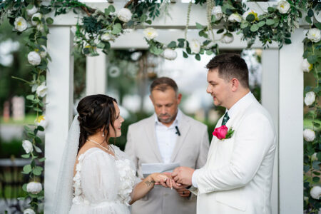 Bride and groom exchange rings under a floral arch at their outdoor wedding ceremony, officiant in background.