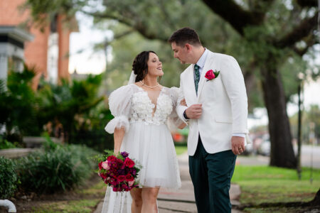 Bride in a short white dress and groom in a white jacket walk arm-in-arm outdoors, smiling amid lush greenery and red flowers.