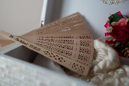 Engraved wooden hand fan in a white box with red and pink faux flowers and white fabric—wedding ceremony décor idea.